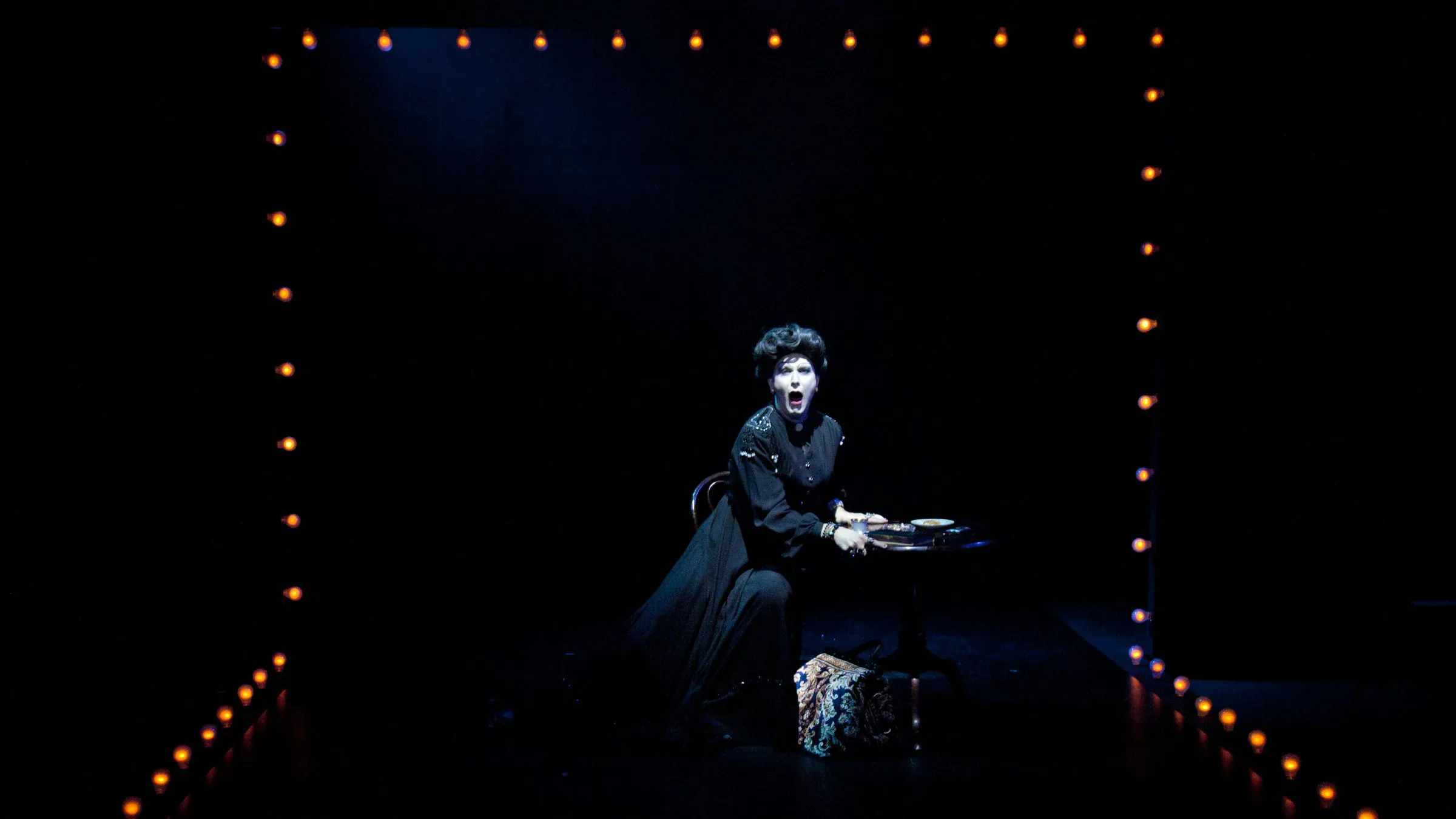 Peter Clements as Frau Welt at a table framed by stage lights