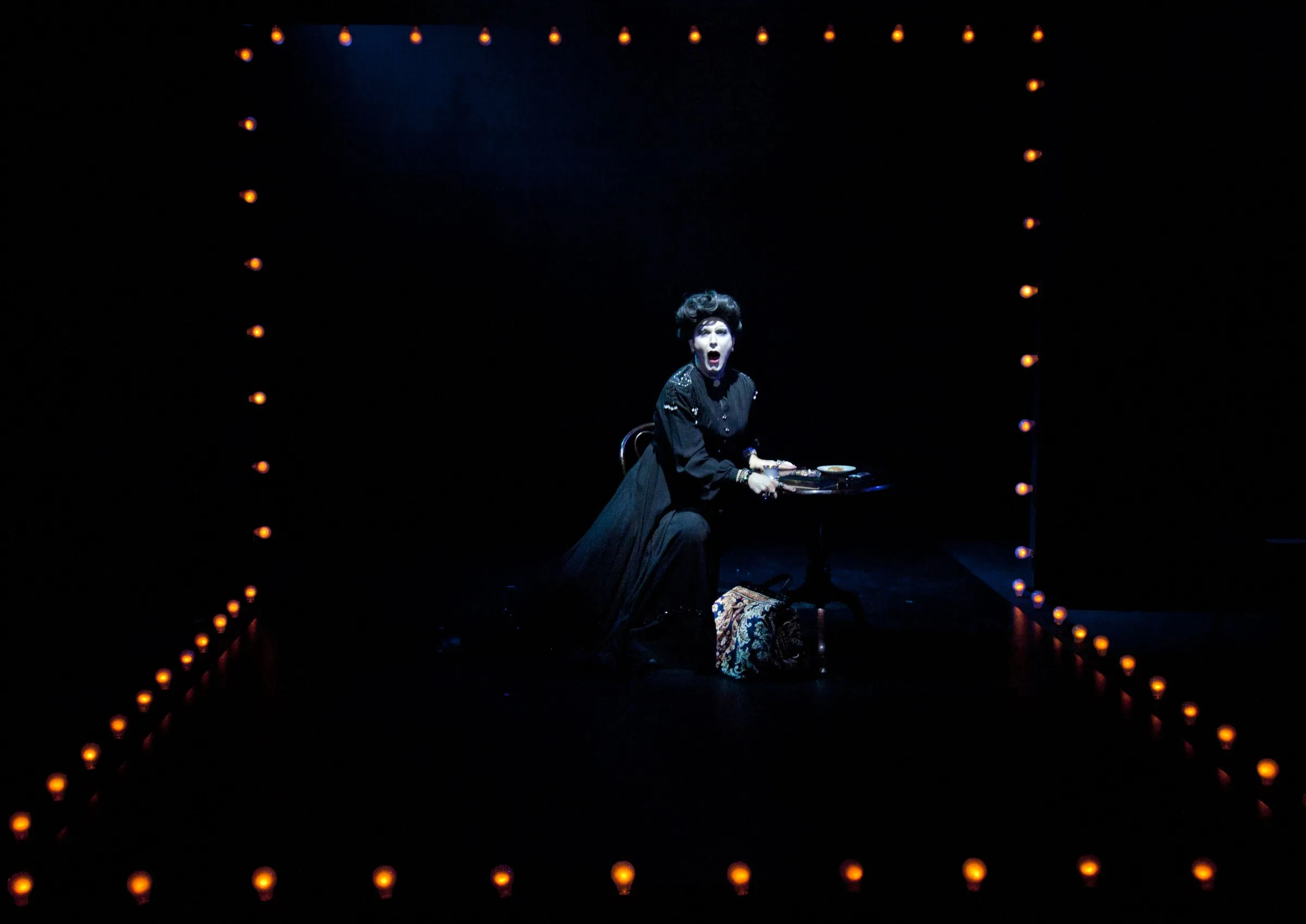 Peter Clements as Frau Welt at a table framed by stage lights