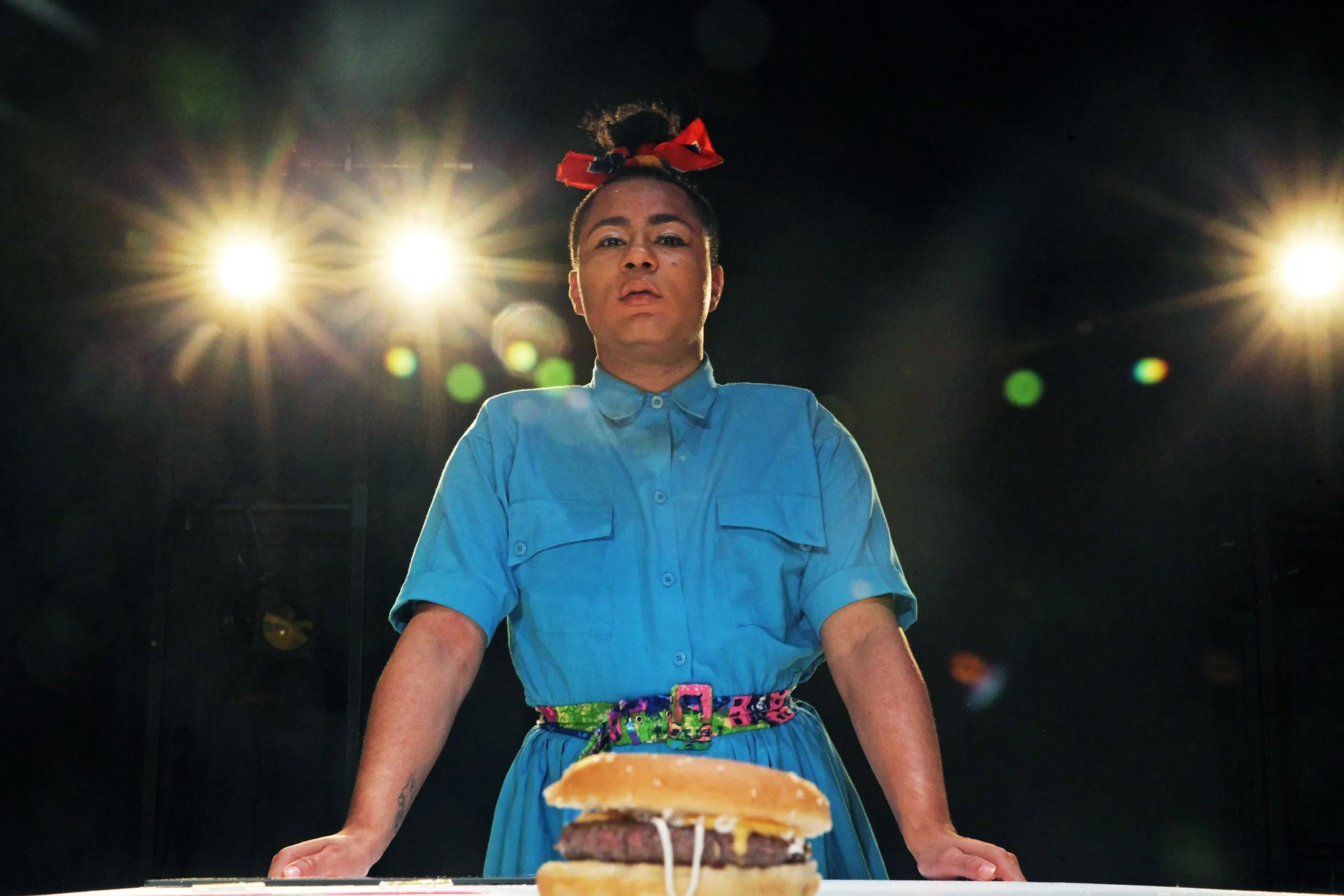 Travis Alabanza stands over a burger under stage lights