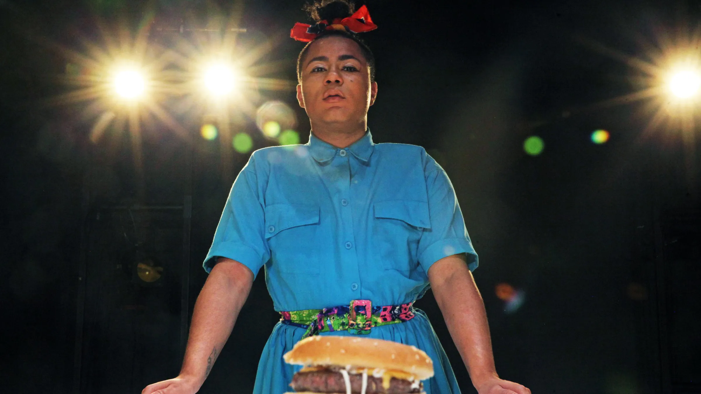 Travis Alabanza stands over a burger under stage lights