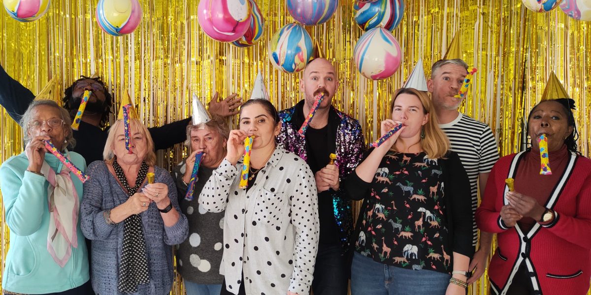 Community members and team members from Hackney Showroom stand in front of a gold sparkly background wearing party hats. We are using this image to promote our community fundraiser, can you help?